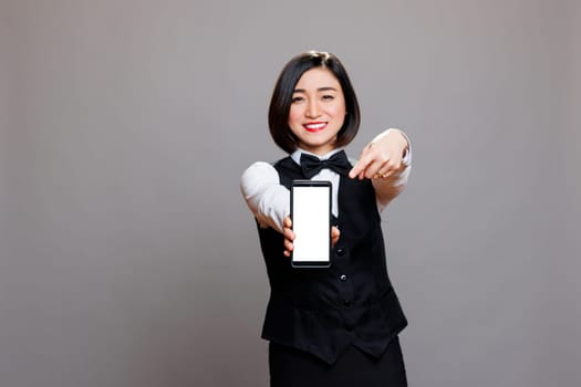 Smiling asian waitress holding smartphone and pointing with finger at blank white screen mockup and looking at camera. Receptionist showing mobile phone blank touchscreen portrait