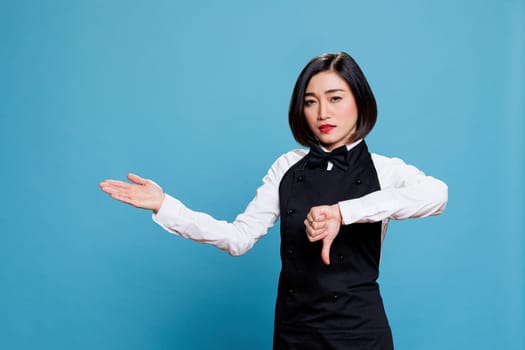 Disappointed asian waitress showing thumb down while pointing to side with arm portrait. Cafe woman employee symbolizing catering service dissatisfaction and showing to right direction