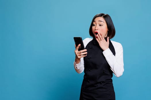 Excited asian woman receptionist covering mouth with surprised gesture while reading client positive feedback on smartphone. Cafe shocked waitress receiving good news on mobile phone