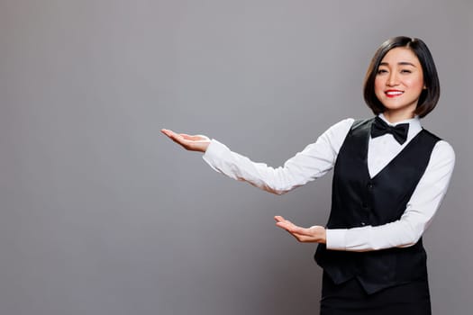 Attractive smiling asian waitress pointing with hands to side and looking at camera. Cheerful young woman receptionist wearing uniform showing with arms, advertising product