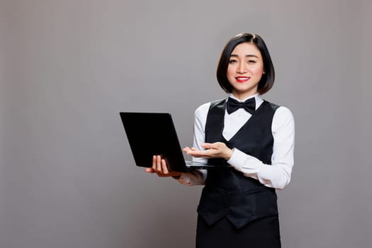 Smiling asian waitress in professional uniform showing laptop and looking at camera. Young attractive receptionist pointing with hand while holding portable computer portrait