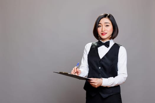Young asian waitress in uniform holding clipboard, writing customer order and looking at camera. Attractive woman receptionist holding pen and checklist studio portrait on gray background