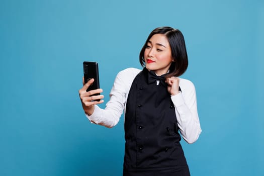 Attractive asian waitress adjusting bow tie and looking at smartphone front camera while taking selfie. Restaurant woman employee in uniform posing making photo on mobile phone