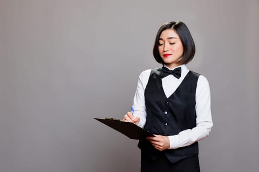 Asian waitress with focused expression writing on clipboard, checking list. Young attractive receptionist dressed in professional black and white uniform taking notes in studio