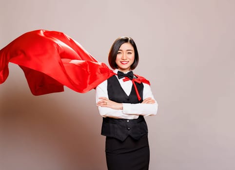 Smiling receptionist standing with folded hands wearing fluttering superwoman cloak, showing confidence and power portrait. Young asian waitress in hero red cape looking at camera