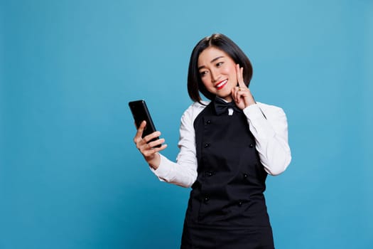 Cheerful asian woman receptionist showing peace gesture with fingers while chatting in smartphone videocall. Smiling young restaurant waitress posing for mobile phone selfie