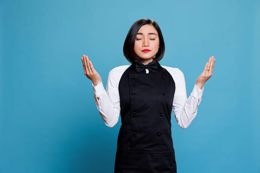 Young attractive asian waitress with closed eyes practicing breathing meditation. Restaurant woman receptionist wearing professional uniform standing with open arms and praying