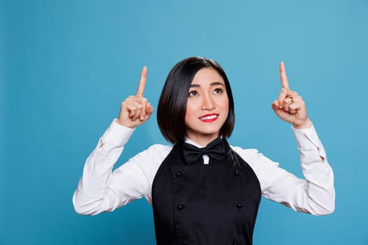 Cheerful smiling asian waitress in uniform pointing with fingers to up direction for catering service advertising. Restaurant woman worker showing upwards for product promotion