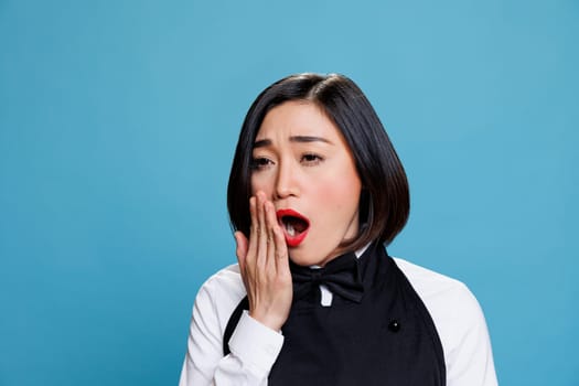 Sleepy young aisan woman receptionist yawning and covering mouth with arm. Restaurant tired overworked waitress wearing uniform feeling exhausted while posing on blue background