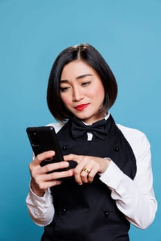 Young smiling asian waitress wearing restaurant uniform tapping on smartphone touchscreen. Attractive woman receptionist answering client message while using mobile phone