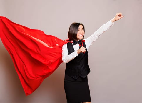 Smiling asian woman receptionist wearing uniform and superman cloak, standing with raised clenched fist. Catering service waitress superhero flying in fluttering red cape while posing in studio