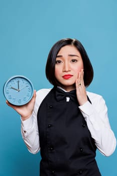 Upset asian waitress showing alarm clock and looking at camera with disappointed facial expression. Sad woman receptionist checking time while running late studio portrait on blue background