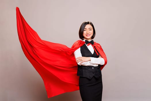 Smiling receptionist standing with folded hands in fluttering superwoman cloak portrait. Attractive young asian woman posing in studio in waitress uniform and superhero cape