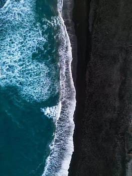 Aerial view of Icelandic black sand beach featuring incredible atlantic beachfront with pounding waves. Cold scene in the north with snow capped mountains and sandy shores with black sand.