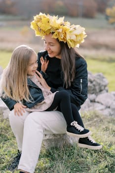 Smiling mother in a wreath of yellow leaves tickles a laughing little girl sitting on her lap on the lawn. High quality photo