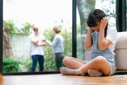 Stressed and unhappy young girl huddle in corner, cover her ears blocking sound of her parent arguing in background. Domestic violence at home and traumatic childhood develop to depression. Synchronos