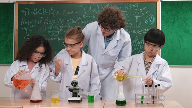 Caucasian smart teacher support excited curious student pour colored liquid in beaker at laboratory. Creative children concentrate to do experiment and enjoy inspecting biochemical sample. Pedagogy.