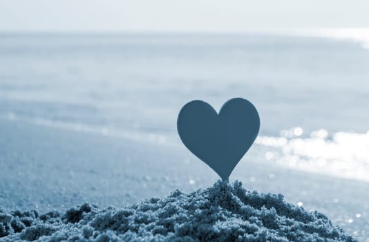 Plastic stick in shape of red heart in sand on sandy beach of sea shore on sea waves on sunny summer day close-up. Concept Love relationship romance amorousness amour St. Valentines Day. Blue color