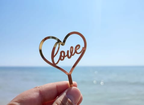 Person holding in fingers hand stick in shape heart and words Love you on background sea and sea waves, blue sky in seashore on sunny summer day close-up. Concept love romance amour St Valentines Day