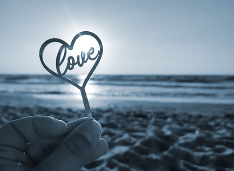 Person holding in fingers hand stick in shape heart and word Love on background sea, waves, blue sky in seashore on sunny summer day close-up. Concept love romance amour St Valentines Day. Blue color