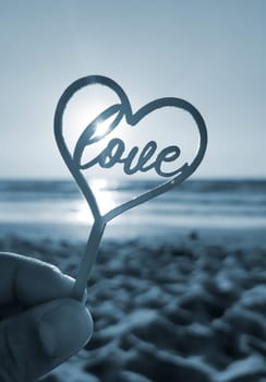 Person holding in fingers hand stick in shape heart and word Love on background sea, waves, blue sky in seashore on sunny summer day close-up. Concept love romance amour St Valentines Day. Blue color