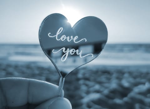 Person holding in fingers hand stick in shape heart and words Love you on sea, waves, sky, sandy beach seashore on sunny summer day close-up. Concept love romance amour St Valentines Day. Blue color