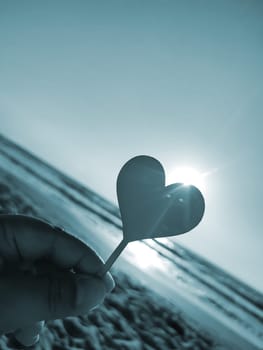 Person holding in fingers hand stick in shape heart on background sea and sea waves, blue sky in seashore on sunny summer day close-up. Concept love romance amour St Valentines Day. Blue color