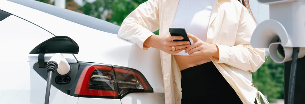 Young woman use smartphone to pay for electricity at public EV car charging station green city park. Modern environmental and sustainable urban lifestyle with EV vehicle. Panorama Expedient