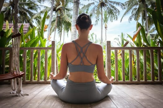 Young woman doing meditation in wooden porch in vacation summer resort. Rear view of young female meditating outdoors. Healthy lifestyle and mental health concept.