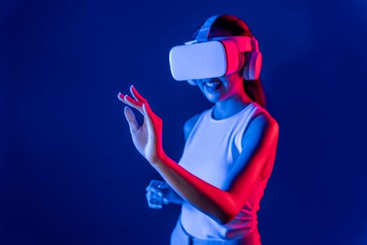 Smart female standing with surrounded by cyberpunk neon light wear VR headset connecting metaverse, futuristic cyberspace community technology. Woman using hand touching virtual object. Hallucination.