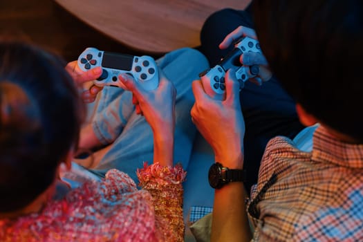 Cropped of hands couple with joysticks sitting sofa playing video game together at back side view with fronted snack and drinks with fun mood at neon light color living room at modern home. Infobahn.