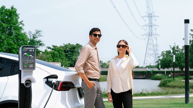 Young couple recharge EV car battery at charging station connected to power grid tower electrical industrial facility as electrical industry for eco friendly vehicle utilization. Expedient