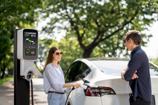 Lovely young couple wearing sun glasses recharging battery for electric car during road trip travel EV car in natural forest or national park. Eco friendly travel during vacation and holiday. Exalt