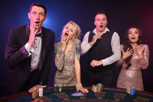Group of four wondered colleagues are playing poker at casino. Youth are making bets waiting for a big win. They are posing standing at the table against a red and blue backlights on black background. Cards, chips, money, gambling, entertainment concept.