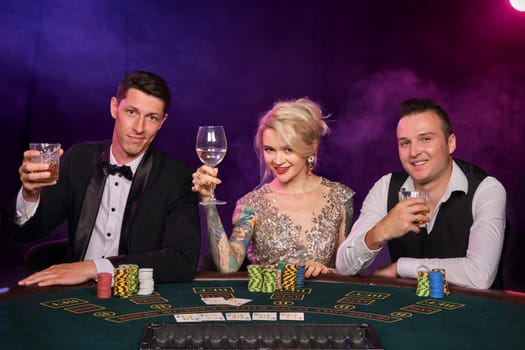 Two stylish men and cute maiden are playing poker at casino. Youth are making bets waiting for a big win. They are smiling and posing sitting at the table against a red and blue backlights on black smoke background. Cards, chips, money, gambling, entertainment concept.