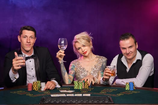 Two stylish fellows and cute lady are playing poker at casino. Youth are making bets waiting for a big win. They are smiling and looking at the camera sitting at the table against a red and blue backlights on black smoke background. Cards, chips, money, gambling, entertainment concept.
