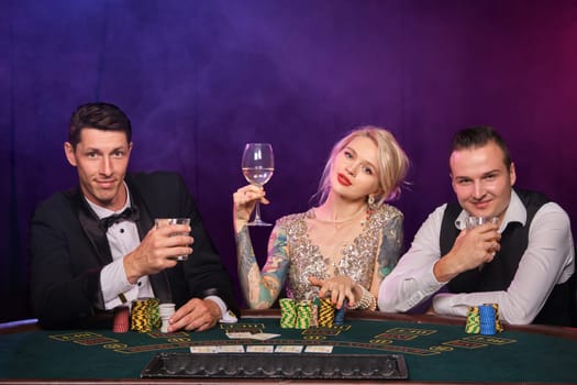 Two stylish guys and cute girl are playing poker at casino. Youth are making bets waiting for a big win. They are smiling and posing sitting at the table against a red and blue backlights on black smoke background. Cards, chips, money, gambling, entertainment concept.