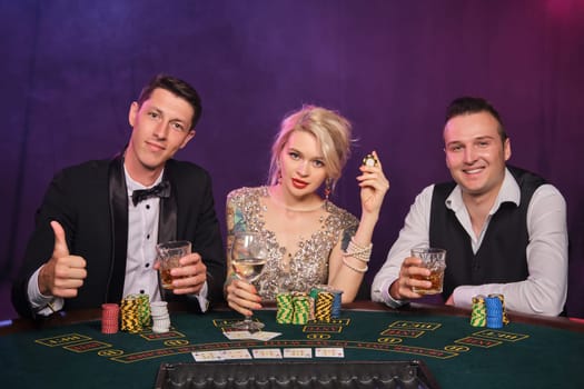Two stylish male and cute female are playing poker at casino. Youth are making bets waiting for a big win. They are smiling and looking at the camera sitting at the table against a red and blue backlights on black smoke background. Cards, chips, money, gambling, entertainment concept.