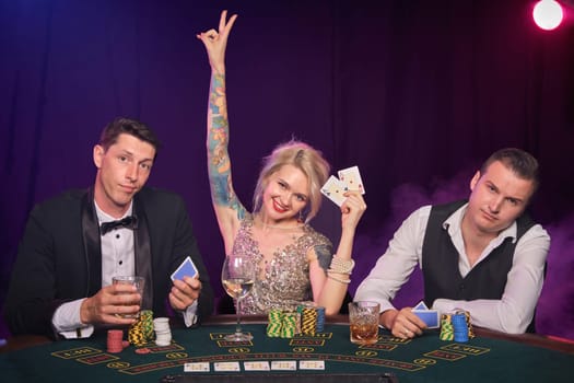 Two stately men and pretty maiden are playing poker at casino. Youth are making bets waiting for a big win. They are smiling and looking at the camera sitting at the table against a red and blue backlights on black smoke background. Cards, chips, money, gambling, entertainment concept.