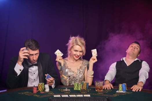 Two stately fellows and pretty lady are playing poker at casino. Youth are making bets waiting for a big win. They posing sitting at the table against a red and blue backlights on black smoke background. Cards, chips, money, gambling, entertainment concept.
