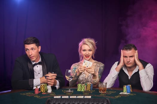 Two stately guys and pretty girl are playing poker at casino. Youth are making bets waiting for a big win. They are smiling and posing sitting at the table against a red and blue backlights on black smoke background. Cards, chips, money, gambling, entertainment concept.