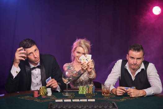 Two stately men and pretty woman are playing poker at casino. Youth are making bets waiting for a big win. They are smiling and looking at the camera sitting at the table against a red and blue backlights on black smoke background. Cards, chips, money, gambling, entertainment concept.