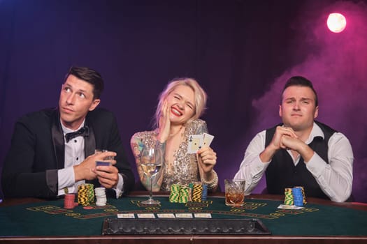 Two stately male and pretty female are playing poker at casino. Youth are making bets waiting for a big win. They are looking upset and posing while sitting at the table against a red and blue backlights on black smoke background. Cards, chips, money, gambling, entertainment concept.