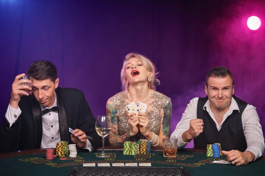 Two rich men and elegant maiden are playing poker at casino. Youth are making bets waiting for a big win. They are posing sitting at the table against a red and blue backlights on black smoke background. Cards, chips, money, gambling, entertainment concept.