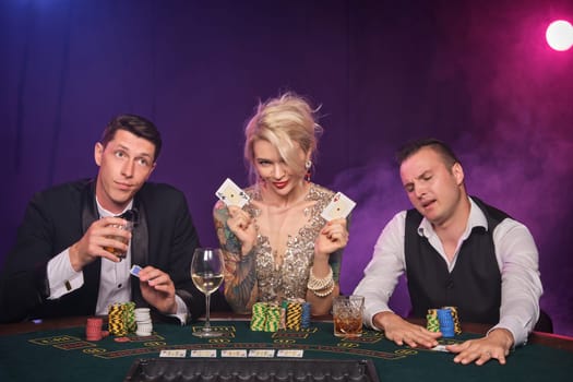 Two rich guys and elegant girl are playing poker at casino. Youth are making bets waiting for a big win. They are posing sitting at the table against a red and blue backlights on black smoke background. Cards, chips, money, gambling, entertainment concept.