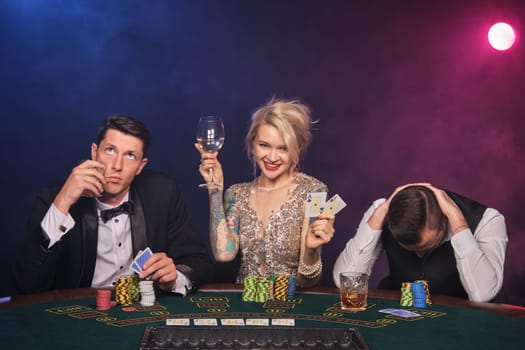 Two rich male and elegant female are playing poker at casino. Youth are making bets waiting for a big win. They are posing sitting at the table against a red and blue backlights on black smoke background. Cards, chips, money, gambling, entertainment concept.