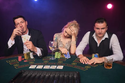 Two wealthy guys and charming girl are playing poker at casino. Youth are making bets waiting for a big win. They are looking disappointed sitting at the table against a red and blue backlights on black smoke background. Cards, chips, money, gambling, entertainment concept.