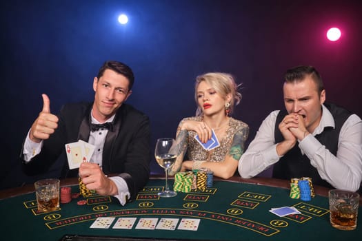 Two wealthy fellows and charming lady are playing poker at casino. Youth are making bets waiting for a big win. They are showing thumb up sitting at the table against a red and blue backlights on black smoke background. Cards, chips, money, gambling, entertainment concept.
