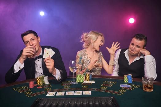 Two wealthy men and charming woman are playing poker at casino. Youth are making bets waiting for a big win. They are looking upset while posing sitting at the table against a red and blue backlights on black smoke background. Cards, chips, money, gambling, entertainment concept.
