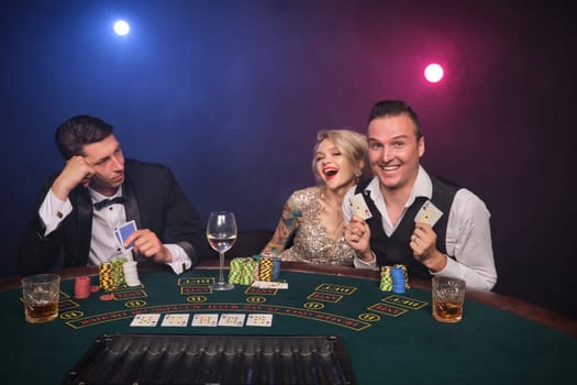 Two athletic men and alluring maiden are playing poker at casino. Youth are making bets waiting for a big win. They are smiling and posing sitting at the table against a red and blue backlights on black smoke background. Cards, chips, money, gambling, entertainment concept.